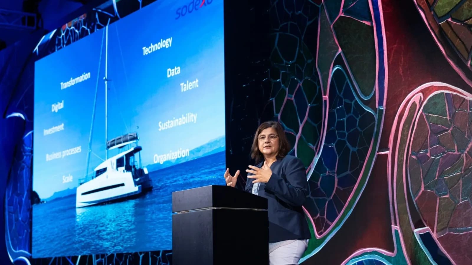 Vera Ingallati speaking from a lectern on a lit stage, a slide mapping transformation, technology, data, talent and sustainability projected behind her.