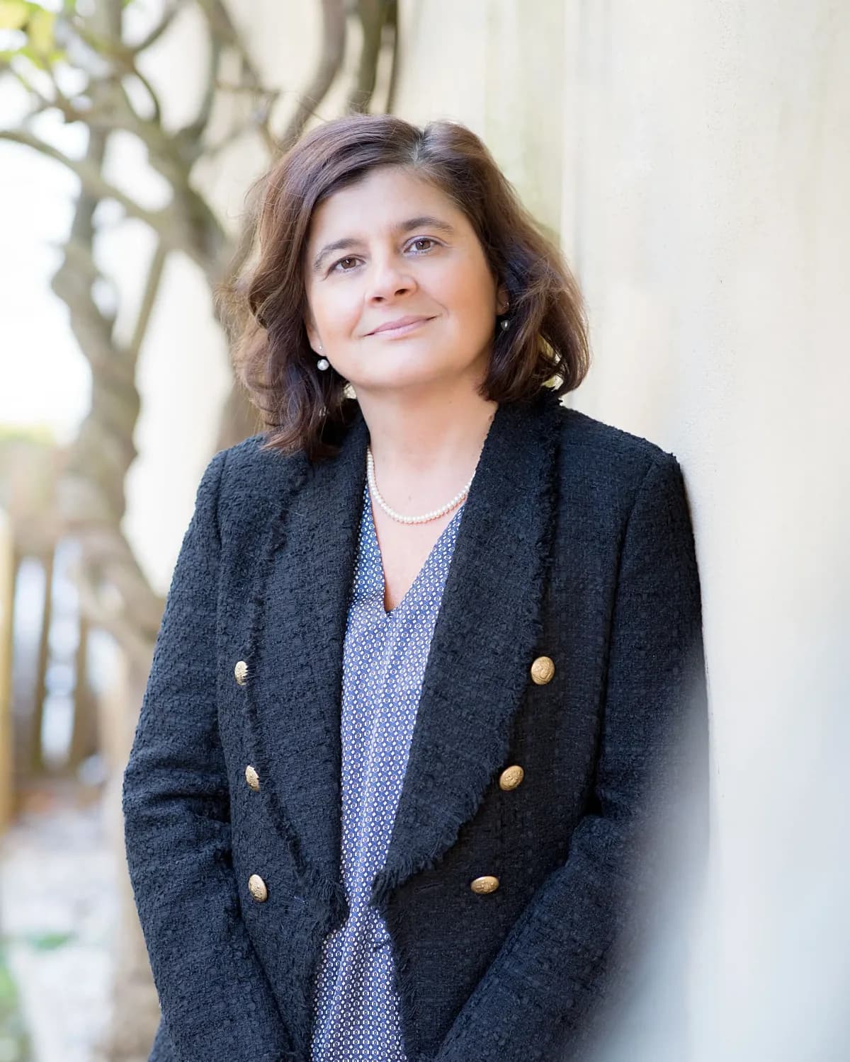 Vera Ingallati outdoors in a dark tweed jacket, leaning against a pale wall with a tree behind her.
