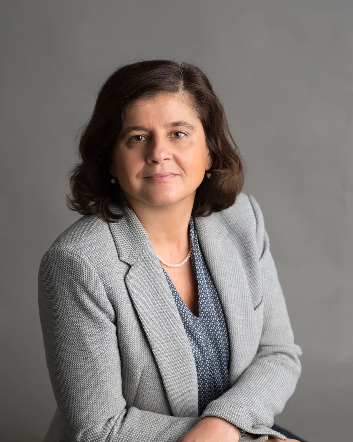 Vera Ingallati seated in a grey jacket, hands resting on the back of a chair, against a plain grey studio wall.